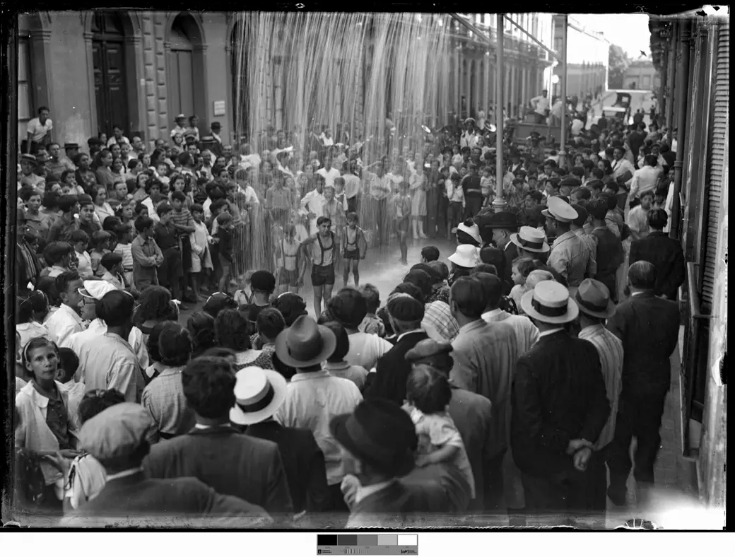 Historical photo, 1940: seen from above, a curtain of water falls onto the middle of a street where a few boys in swimming trunks and shorts stand, hemmed in by a dense crowd in straw hats and caps, with a truck in the distance.