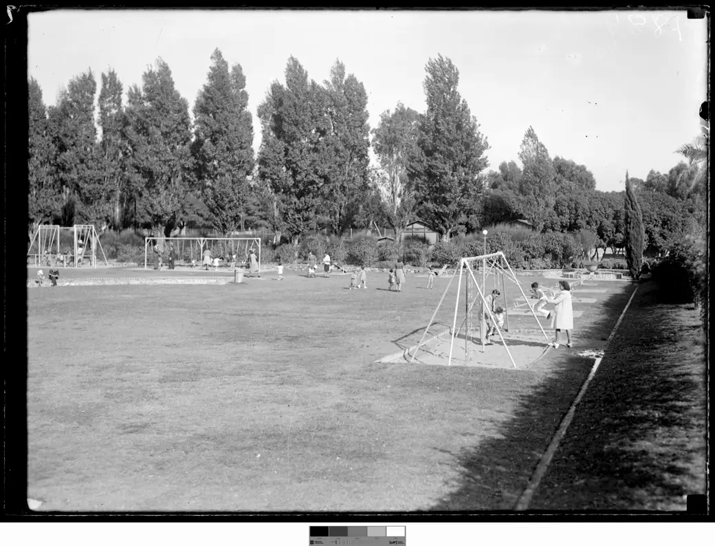 Historical photo, 1942: a wide mown lawn with swing sets, the nearest on sand, a man, a woman and small children beside it, more children and adults further off, and a line of tall trees behind.
