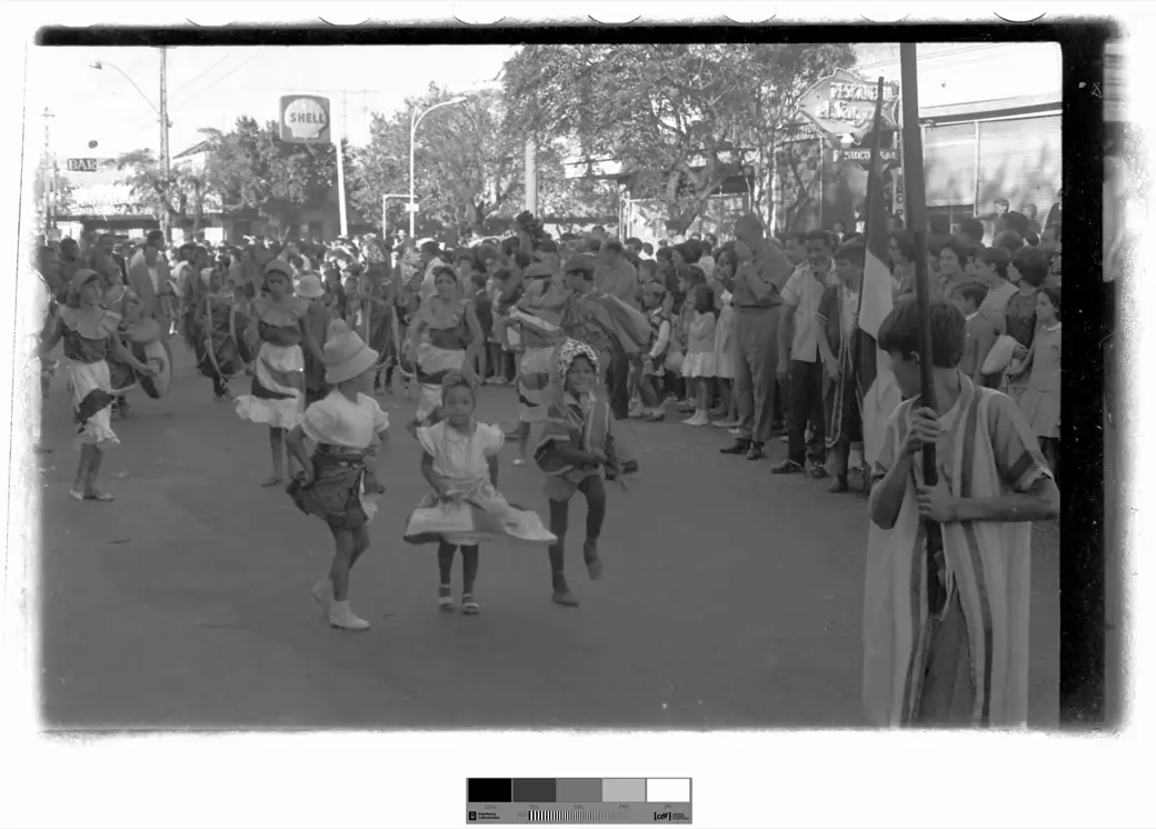 Historical photo, 1966: three small children in costume dance in the middle of a wide street, with a line of costumed dancers behind them, a crowd along the right-hand side and, behind the crowd, a Shell sign on a tall pole.