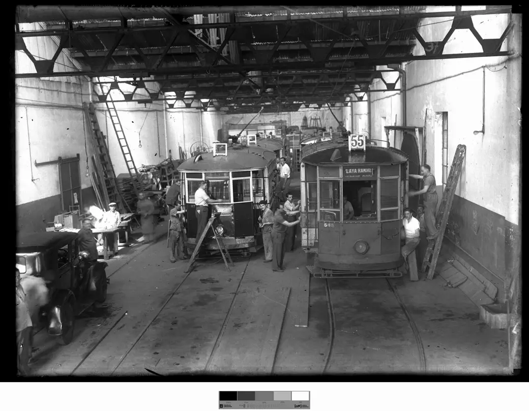 A workshop under a metal-truss roof: tramcars on the rails, two in the foreground, one showing route 55, with workers and ladders around them.