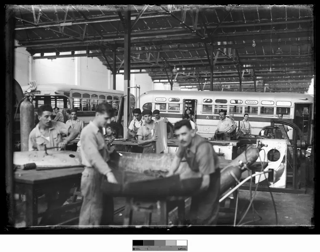 Workers shape a sheet of bodywork metal by hand at a bench; behind them, two buses under the shed roof.
