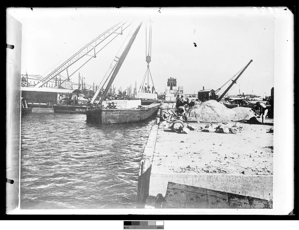 Historical photo, 1901-1909: the port under construction, a crane moving dressed stone from the barge to the quay, a second barge moored alongside.