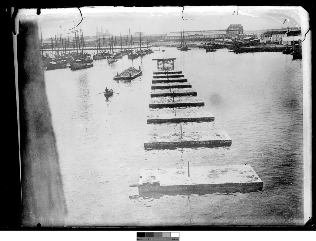Historical photo, 1901-1909: a line of floating concrete caissons queued toward the works of dársena 2, a rowboat and a laden barge nearby.
